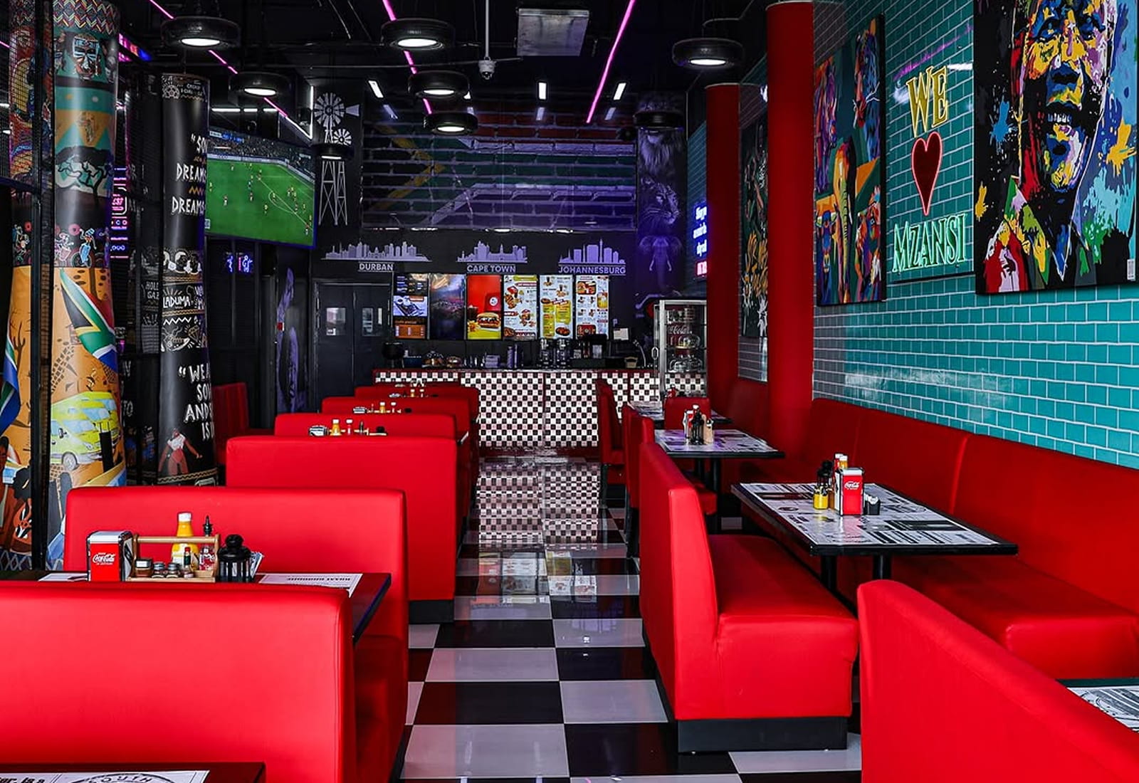 A vibrant restaurant interior featuring bright red booth seating, black-and-white checkered flooring, colorful wall art, and a counter area decorated with bold graphics and menus.