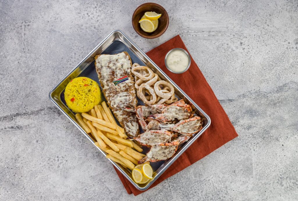 A seafood platter featuring grilled fish topped with a creamy sauce, prawns, fried calamari rings, French fries, and yellow rice, served on a metal tray with lemon wedges and a small cup of dipping sauce.