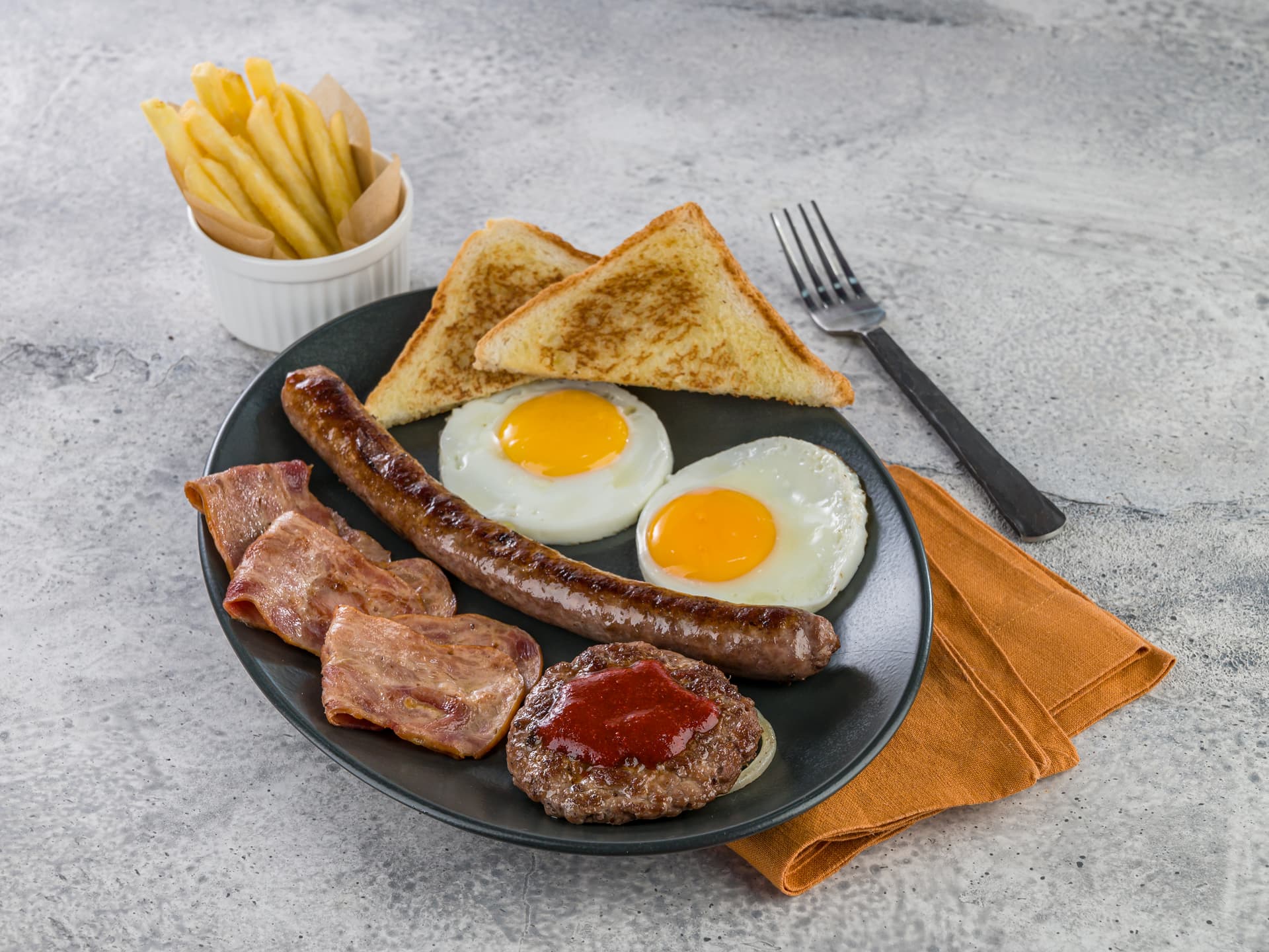 A breakfast plate with two sunny-side-up eggs, toast, sausage, grilled meat slices, and a patty topped with ketchup, served with a side of French fries.