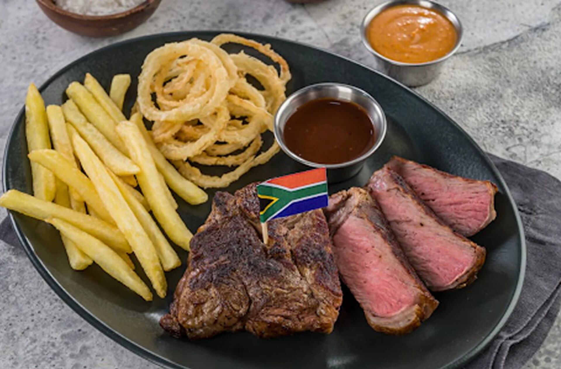 A plate with sliced medium-rare steak, a grilled beef portion topped with a small flag, French fries, crispy onion rings, and a side of dipping sauce.