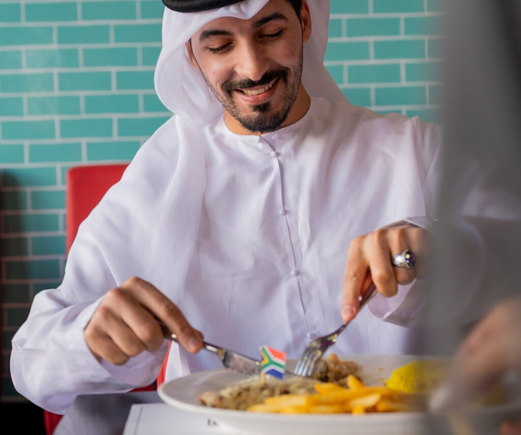 Man in traditional attire enjoying breakfast, adding sauce to his meal in a bright and casual restaurant setting.