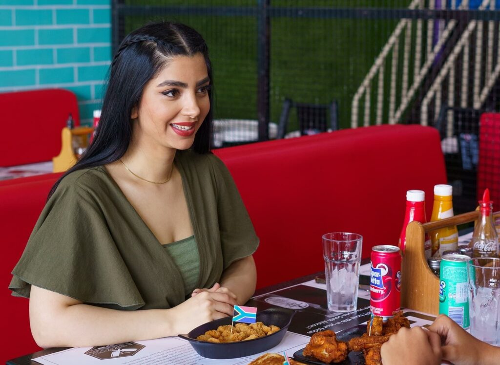 Woman enjoying a casual meal at a colorful restaurant table with drinks and condiments, smiling in a relaxed and friendly dining setting.