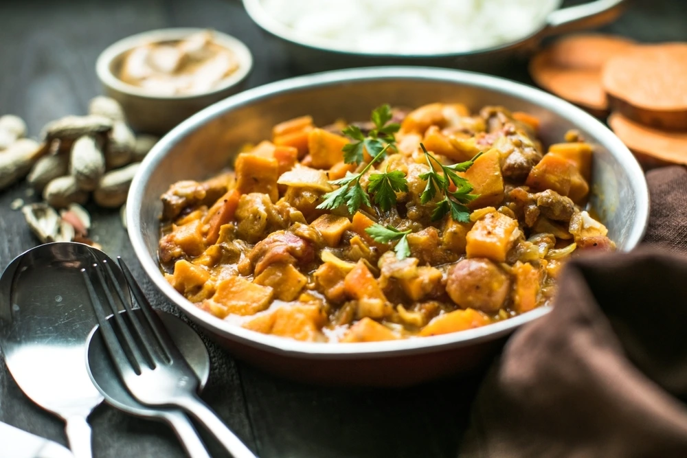 A bowl of South African peanut and sweet potato stew garnished with fresh parsley, served on a rustic table with a spoon and fork beside it, surrounded by peanuts, sliced sweet potatoes, and a small bowl of sauce in the background.