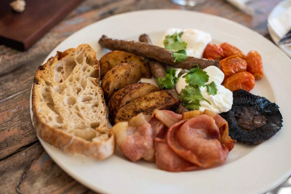 A plate of breakfast food featuring toasted bread slices, roasted potatoes, bacon, sausages, poached eggs topped with herbs, roasted tomatoes, and a grilled mushroom.