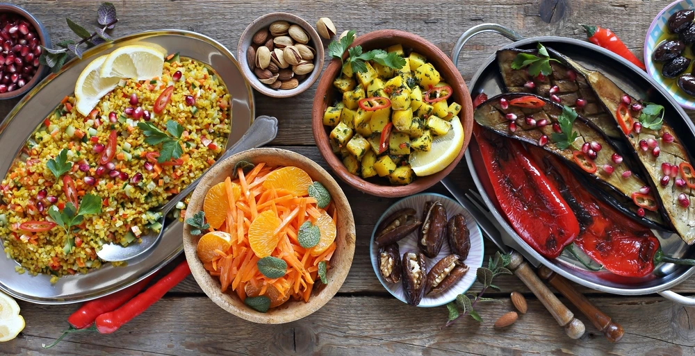 A colorful spread of Middle Eastern–style dishes on a rustic wooden table, including bulgur salad with pomegranate, roasted eggplant and peppers, seasoned potatoes, a carrot and orange salad, stuffed dates, and bowls of nuts and olives.
