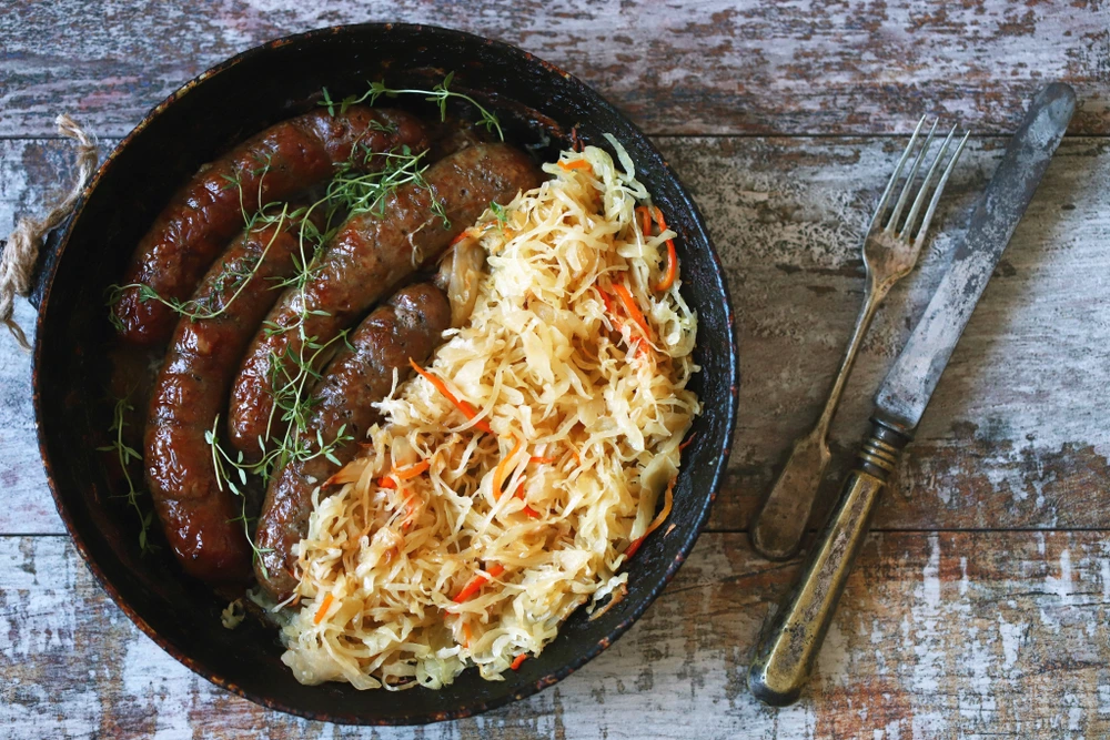 A rustic pan filled with browned sausages and a generous serving of sauerkraut with shredded carrots, placed on a weathered wooden surface alongside a vintage fork and knife.
