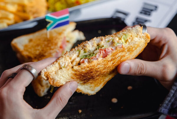 Hands holding a toasted sandwich filled with a creamy mix of chopped vegetables, with another sandwich piece on a plate behind it topped with a small South African flag