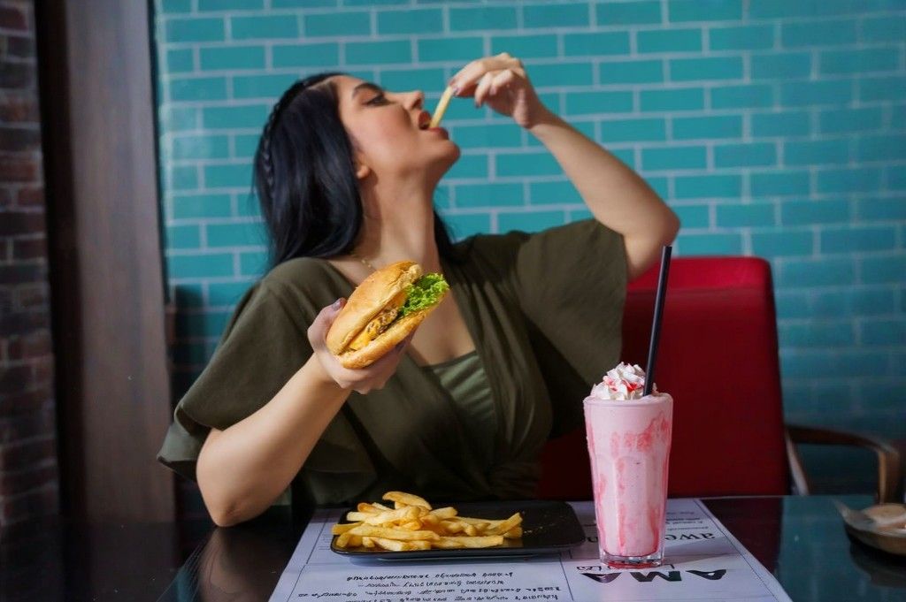 Woman enjoying a burger and fries with a pink milkshake while dining at a modern restaurant.