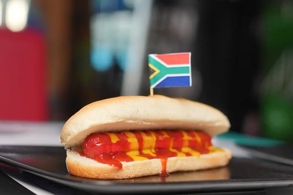 Hot dog with ketchup and mustard, topped with a South African flag, served on a black plate.
