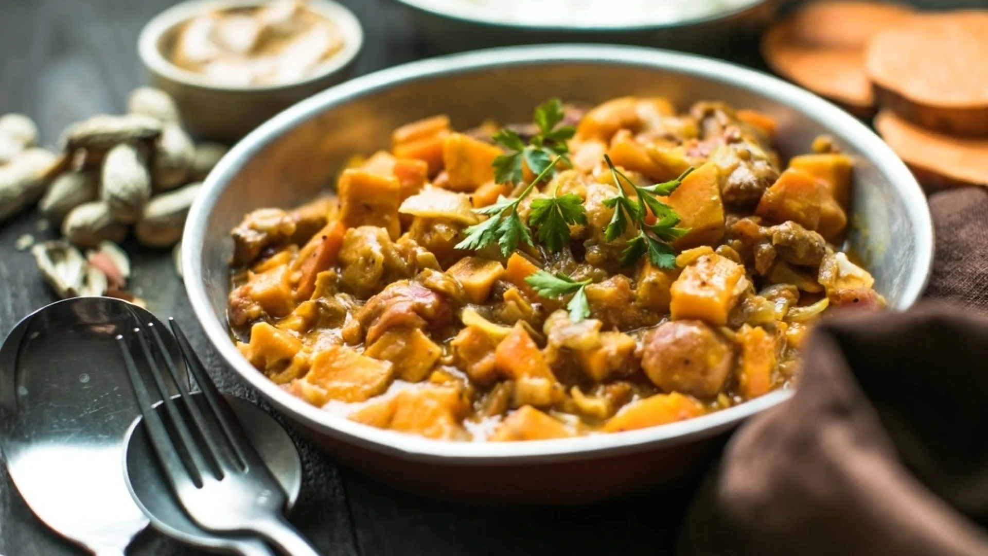 A bowl of South African peanut and sweet potato stew garnished with fresh parsley, served on a rustic table with a spoon and fork beside it, surrounded by peanuts, sliced sweet potatoes, and a small bowl of sauce in the background.