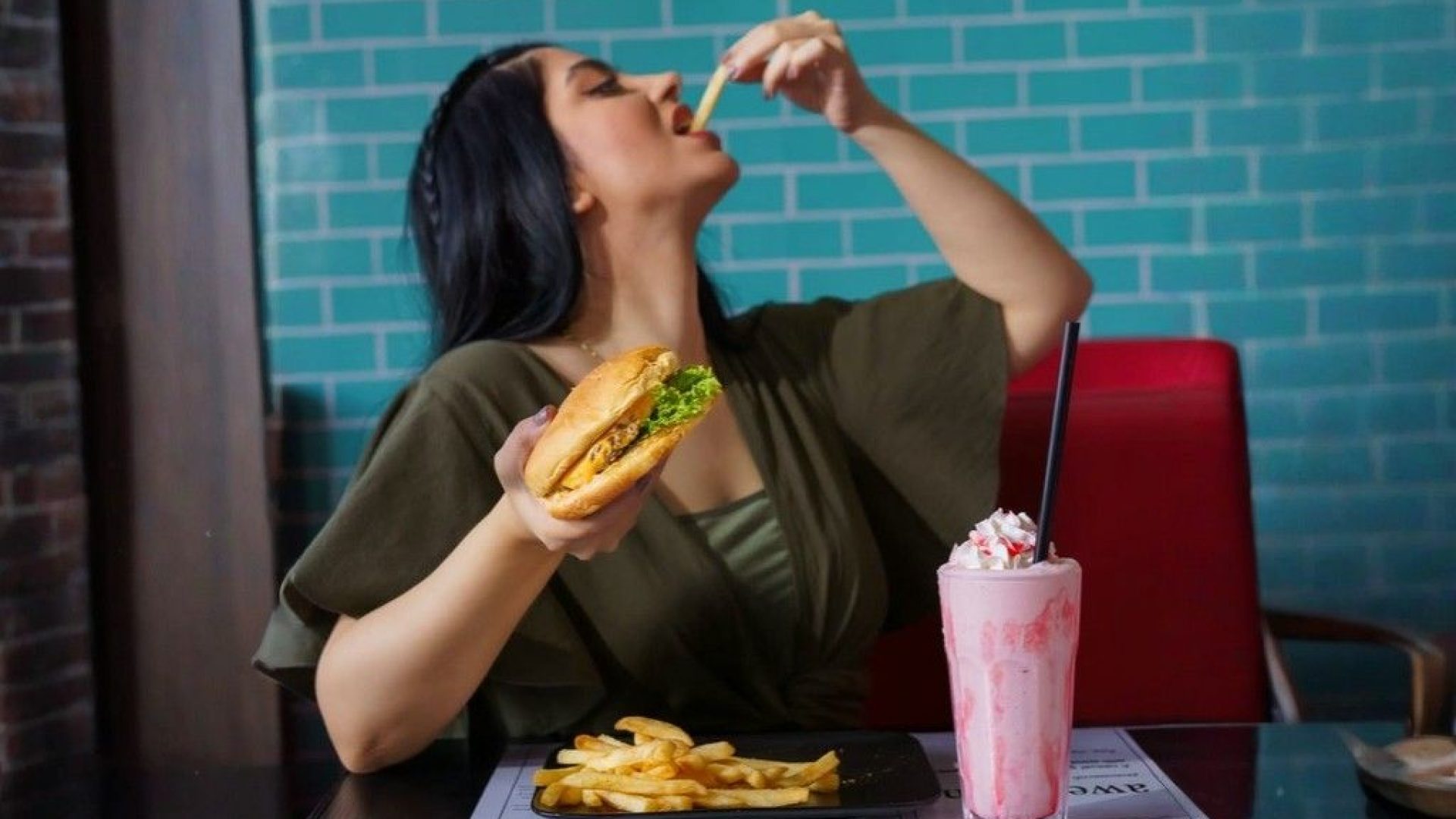 Woman enjoying a burger and fries with a pink milkshake while dining at a modern restaurant.