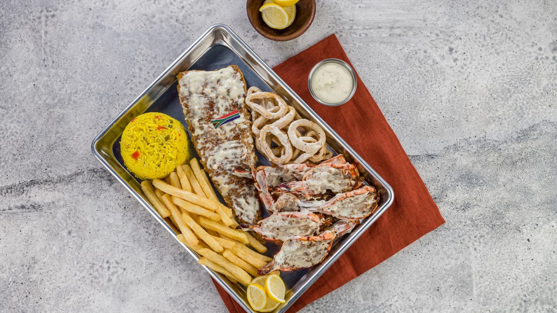 A seafood platter featuring grilled fish topped with a creamy sauce, prawns, fried calamari rings, French fries, and yellow rice, served on a metal tray with lemon wedges and a small cup of dipping sauce.