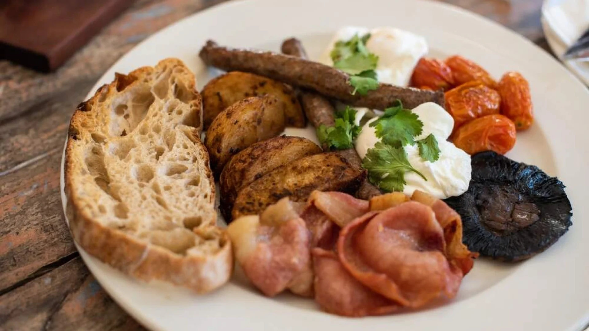 A plate of breakfast food featuring toasted bread slices, roasted potatoes, bacon, sausages, poached eggs topped with herbs, roasted tomatoes, and a grilled mushroom.