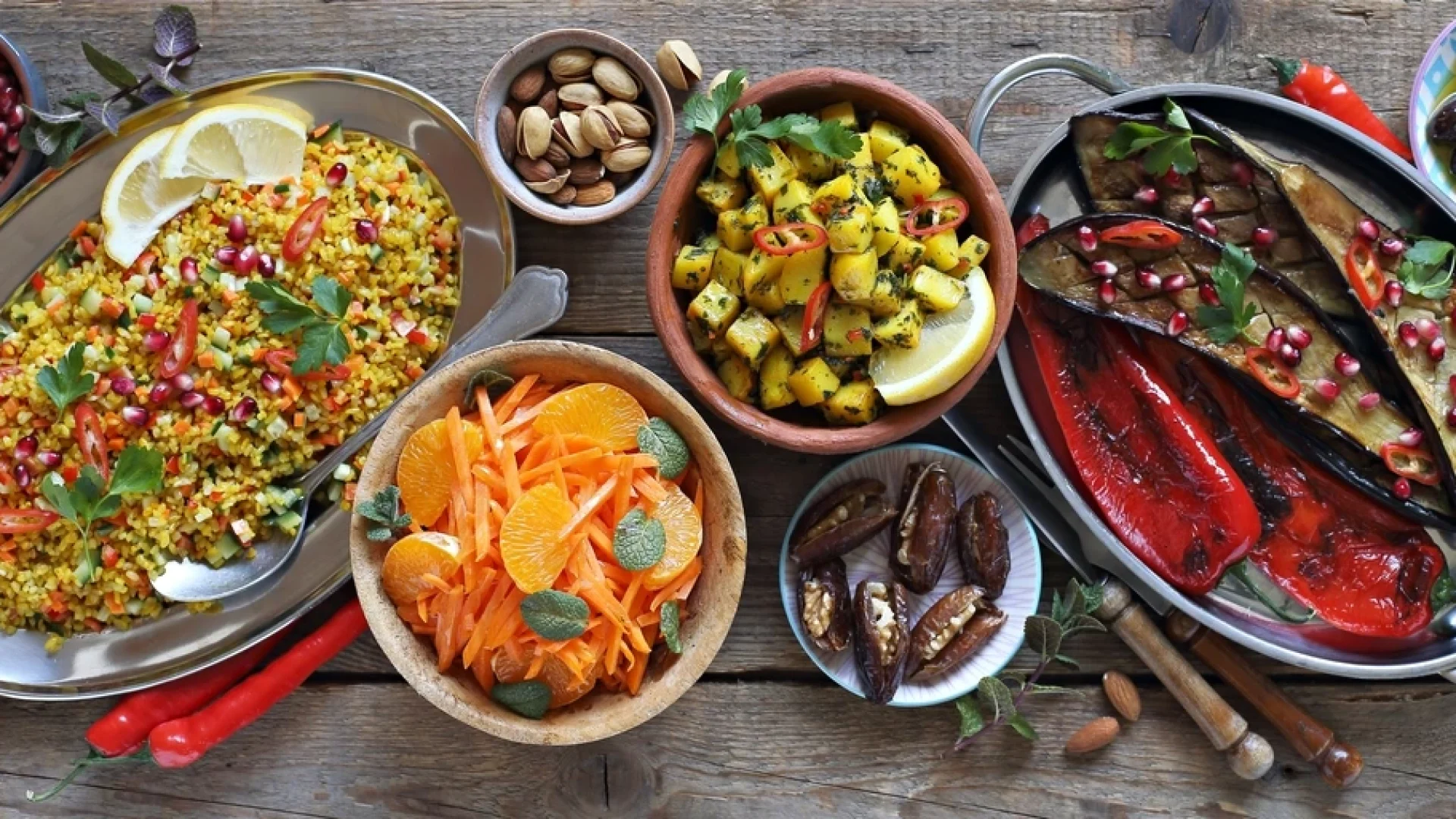 A colorful spread of Middle Eastern–style dishes on a rustic wooden table, including bulgur salad with pomegranate, roasted eggplant and peppers, seasoned potatoes, a carrot and orange salad, stuffed dates, and bowls of nuts and olives.