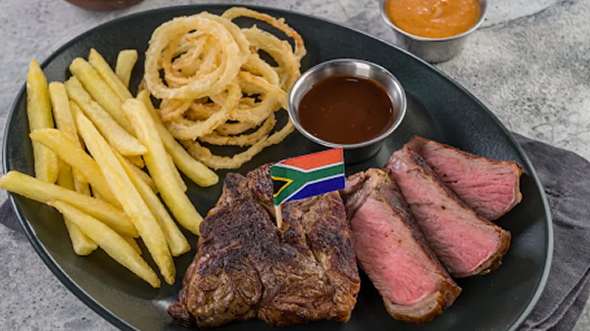 A plate with sliced medium-rare steak, a grilled beef portion topped with a small flag, French fries, crispy onion rings, and a side of dipping sauce.