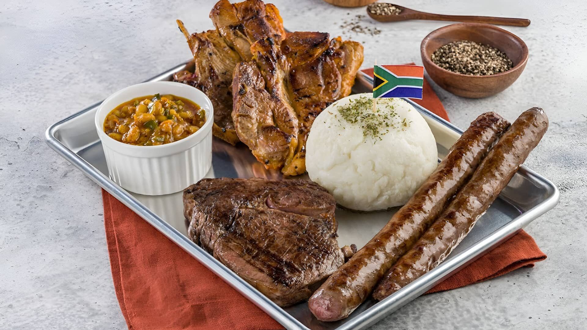 South African platter with grilled steak, boerewors sausage, chicken, pap, and chakalaka served on a tray with traditional flavors.