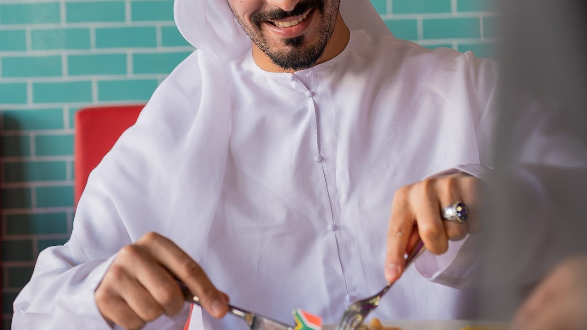 Man in traditional attire enjoying breakfast, adding sauce to his meal in a bright and casual restaurant setting.
