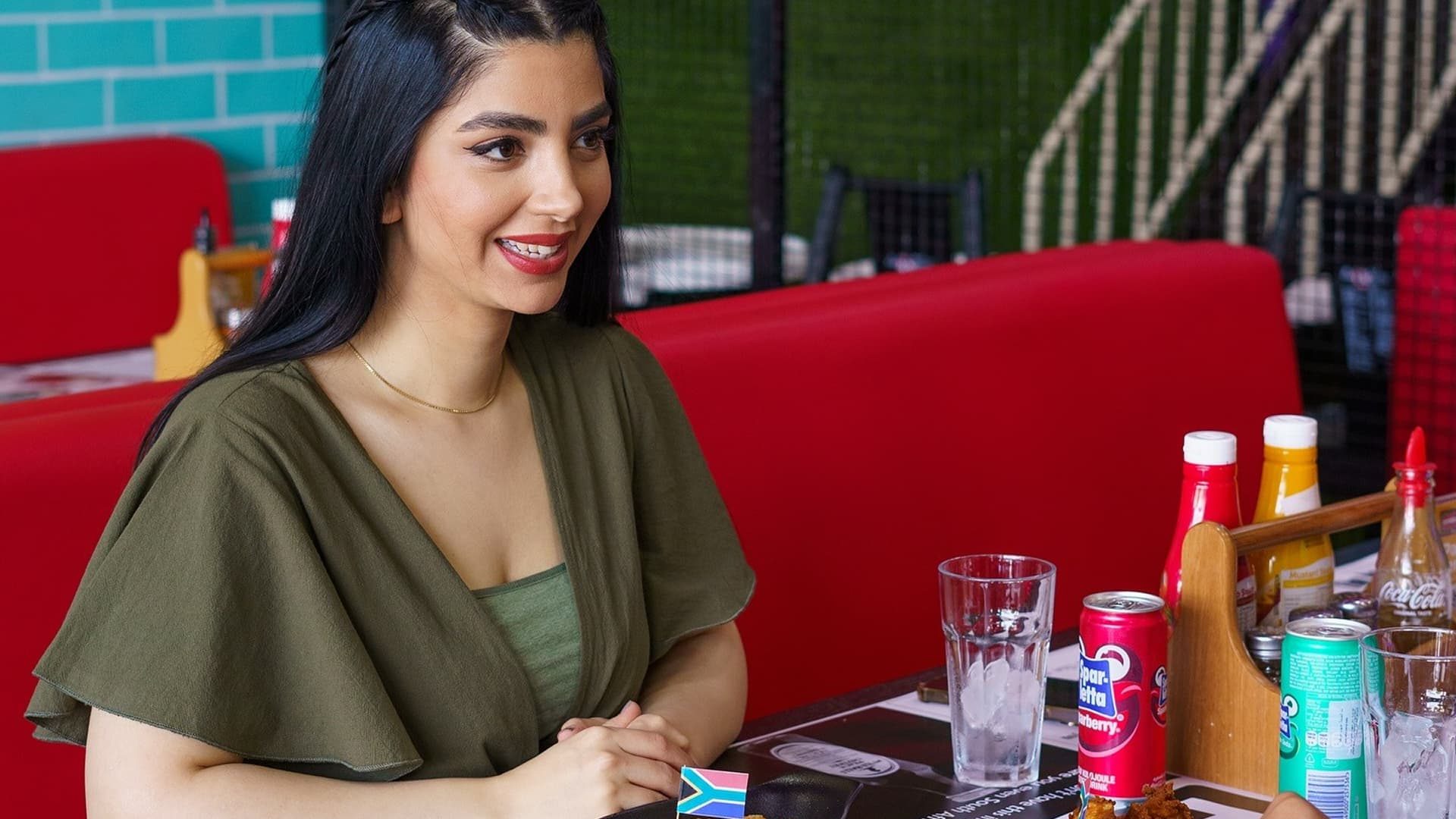Woman enjoying a casual meal at a colorful restaurant table with drinks and condiments, smiling in a relaxed and friendly dining setting.