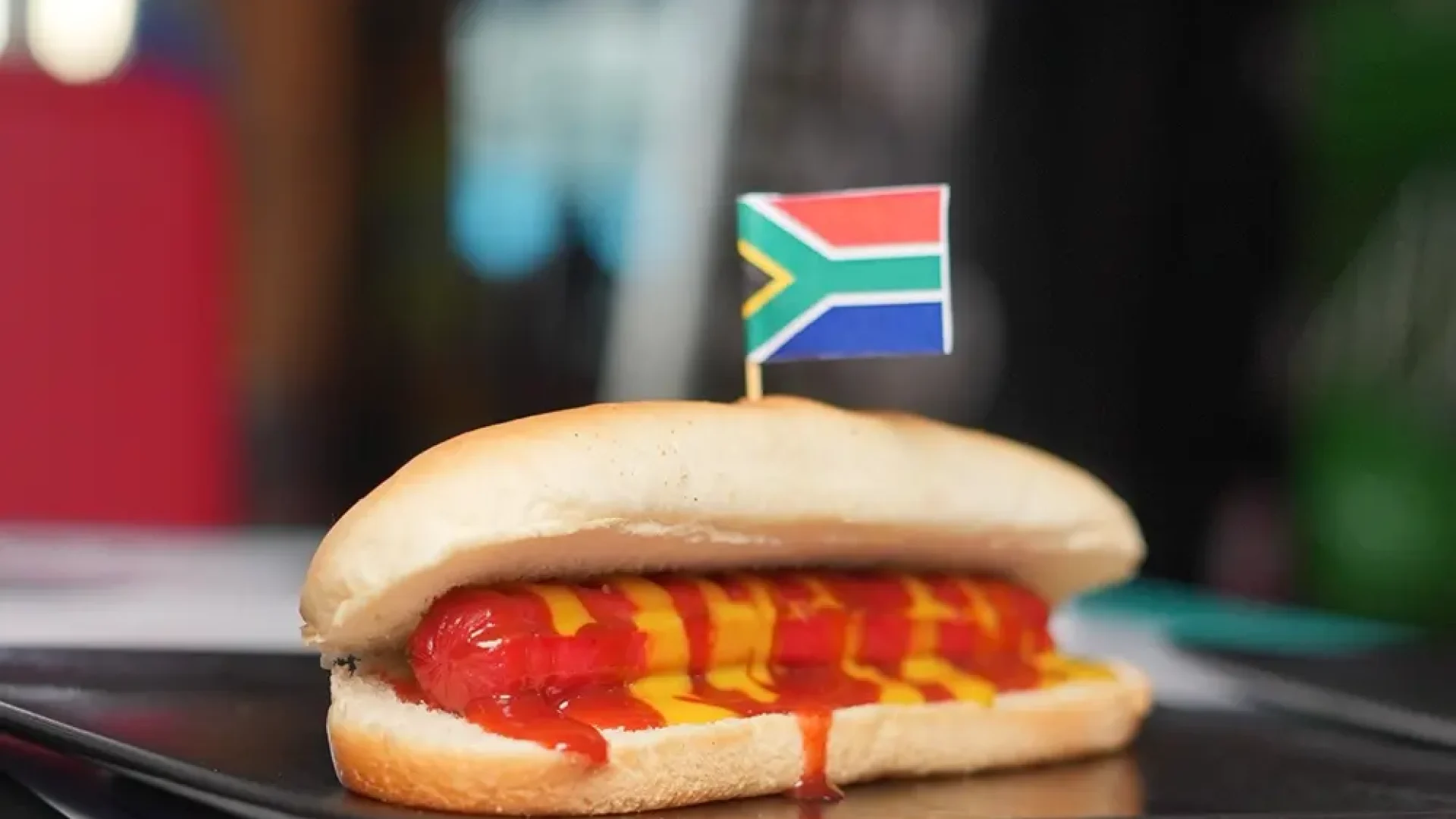 Hot dog with ketchup and mustard, topped with a South African flag, served on a black plate.