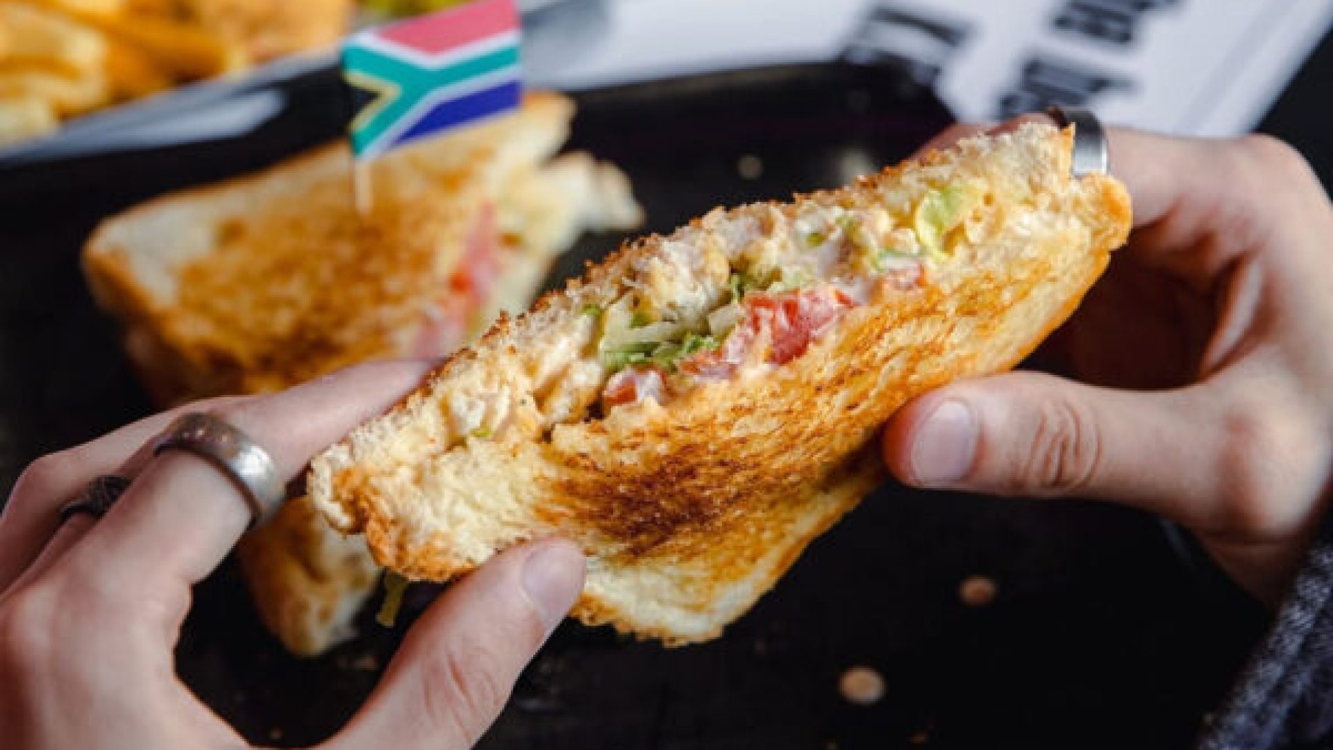 Hands holding a toasted sandwich filled with a creamy mix of chopped vegetables, with another sandwich piece on a plate behind it topped with a small South African flag