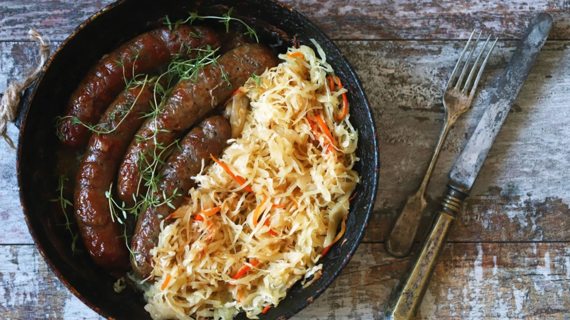 A rustic pan filled with browned sausages and a generous serving of sauerkraut with shredded carrots, placed on a weathered wooden surface alongside a vintage fork and knife.