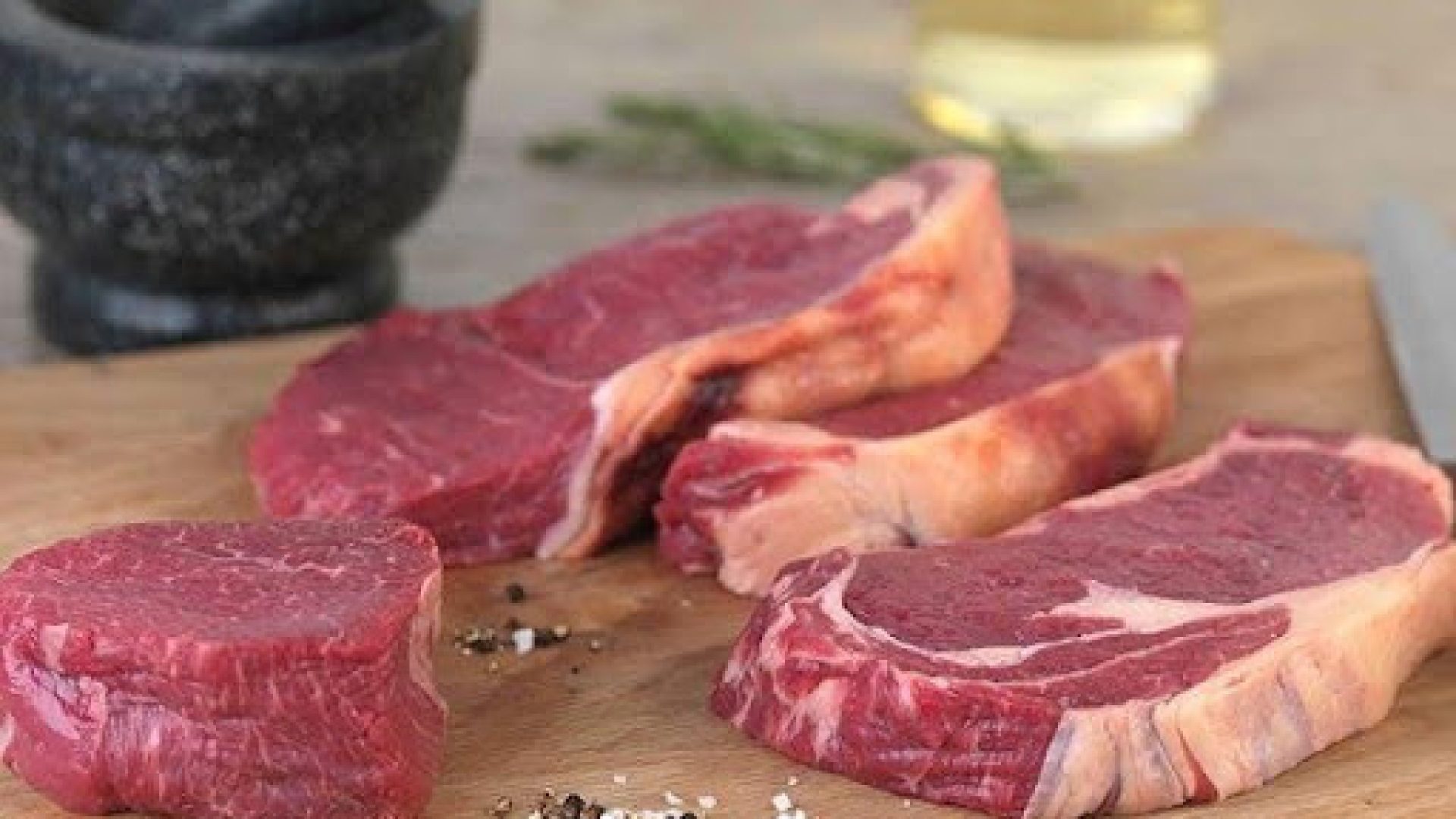 Raw ribeye and sirloin steak cuts on a wooden board with mortar and pestle, rosemary, and olive oil
